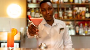 A bartender holding a pink cocktail with an orange garnish. In the background, there are shelves with various bottles. - Forest Hill Hotel (Arusha)