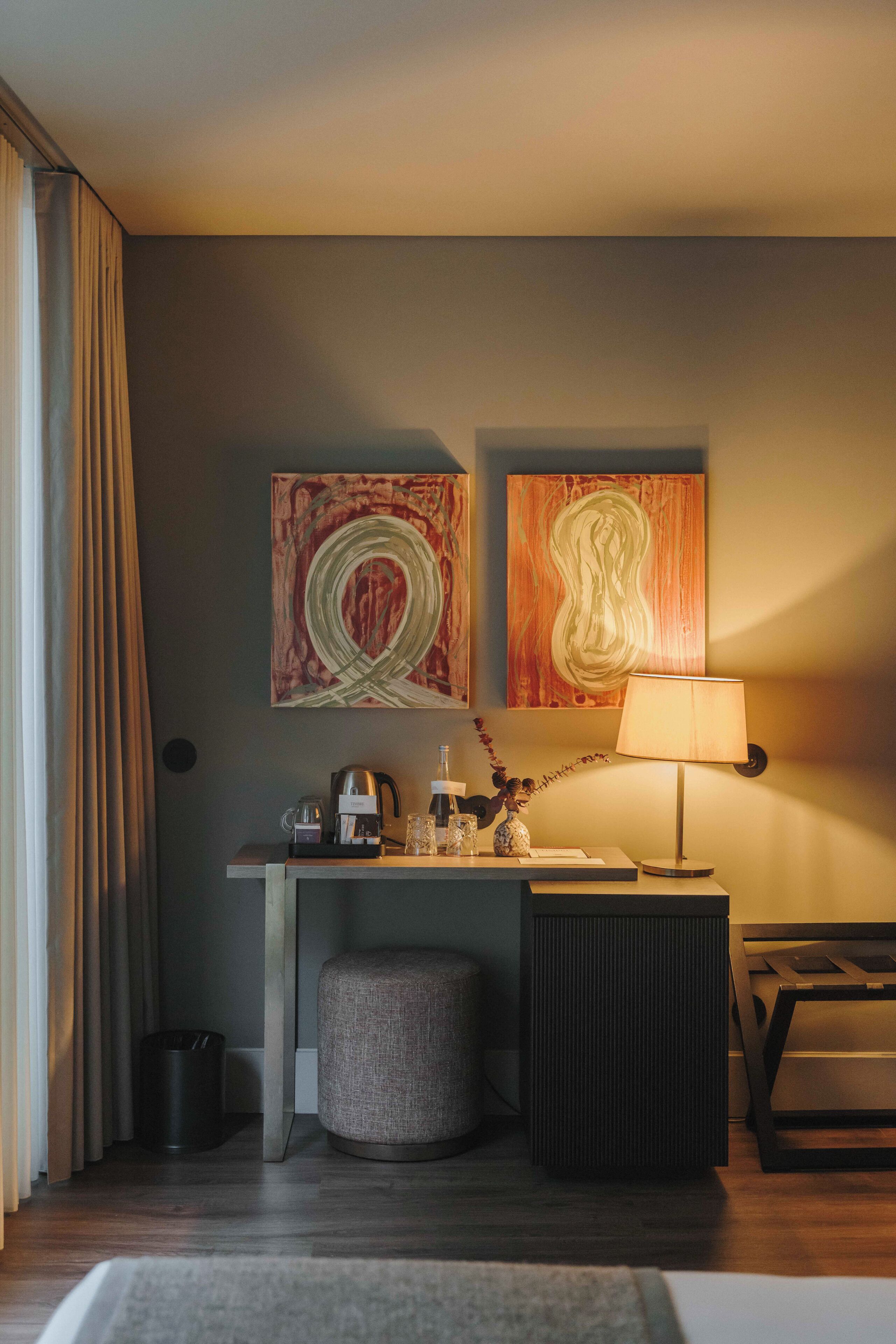 Ein Hotelzimmer mit Schreibtisch, Lampe, zwei abstrakten Gemälden, einem Mülleimer und einem kleinen Hocker.