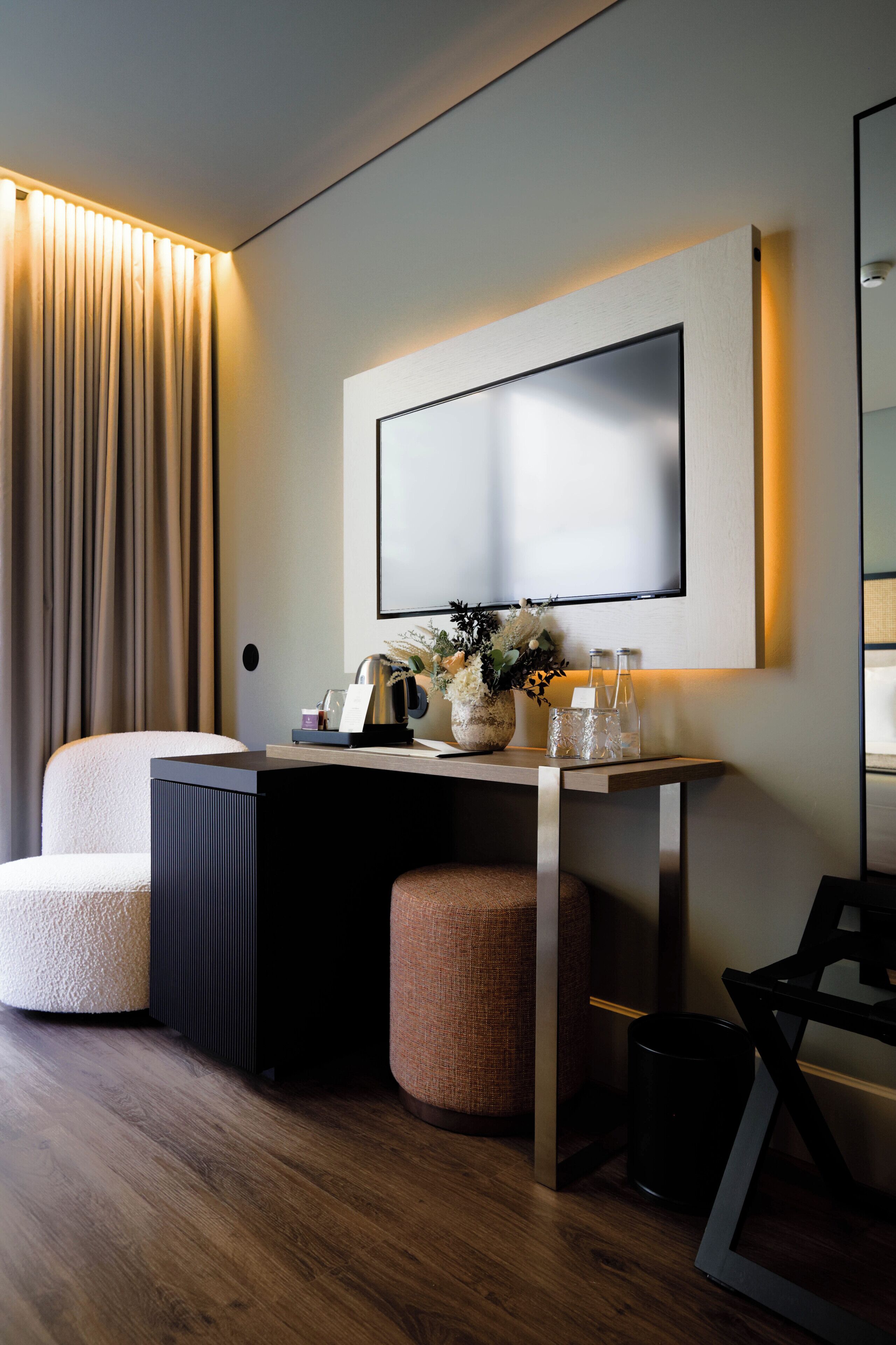 Habitación de hotel moderna con una televisión de pantalla plana grande montada en la pared, un escritorio de madera con un jarrón de flores, una silla y un taburete pequeño.