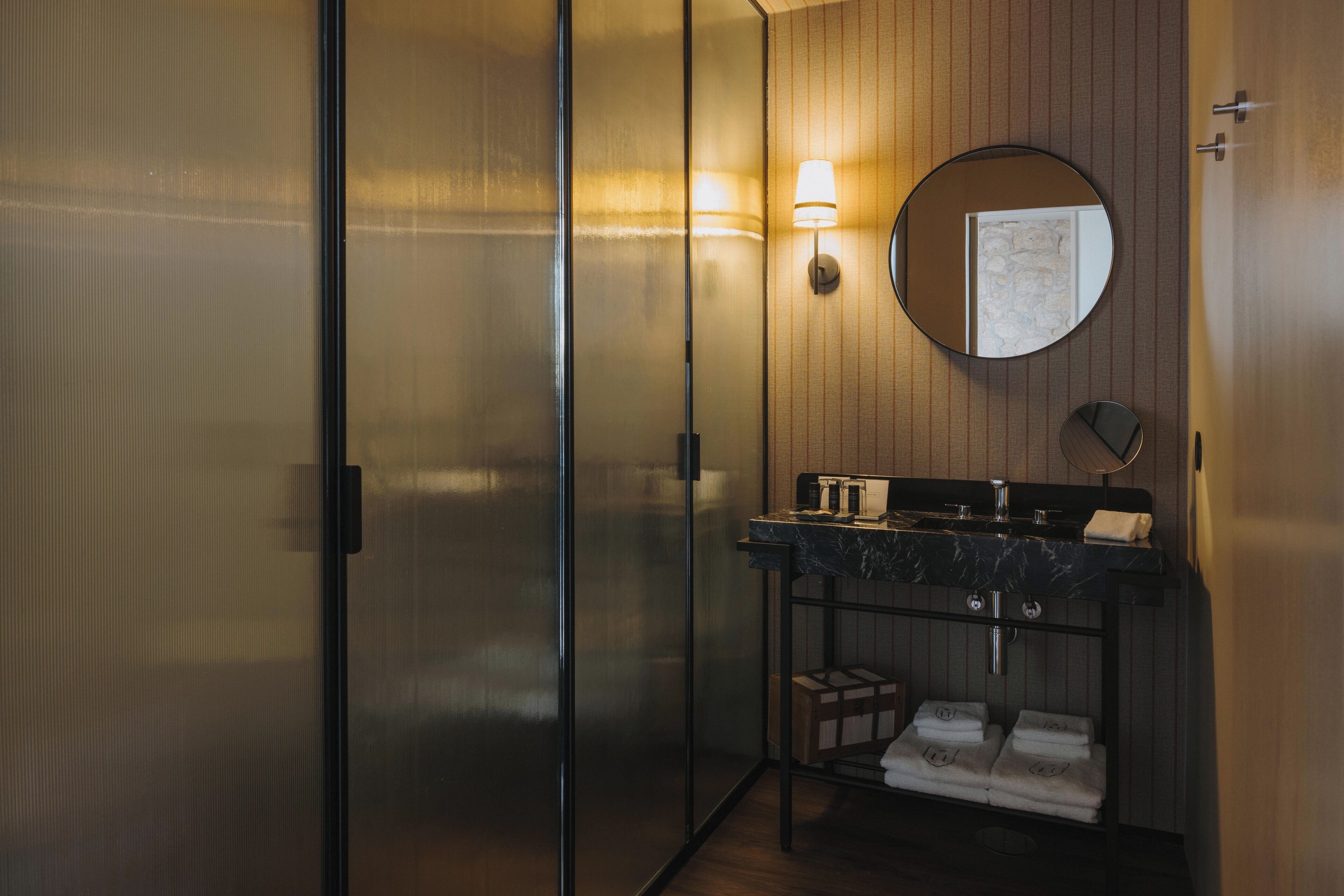 A modern bathroom with a glass shower enclosure, a wooden vanity with a marble countertop, and a round mirror.