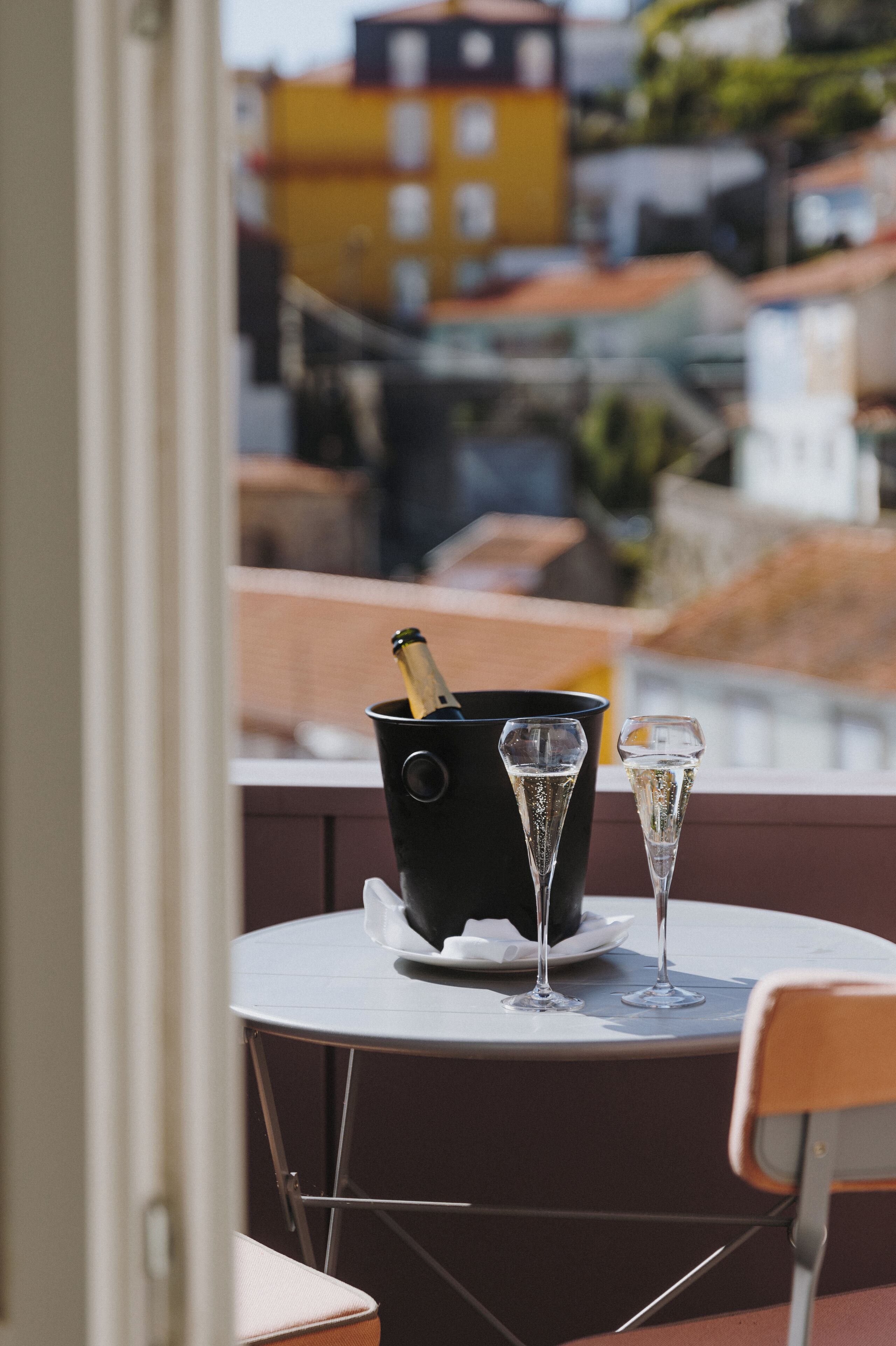 Een tafel met een zwarte emmer, waarin een fles en twee champagneglazen staan, op een balkon met uitzicht op een stadsgezicht.
