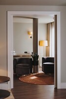 Un vestíbulo de hotel moderno con suelo de madera, dos sillones oscuros, una mesa de centro redonda, una planta en maceta y una lámpara.