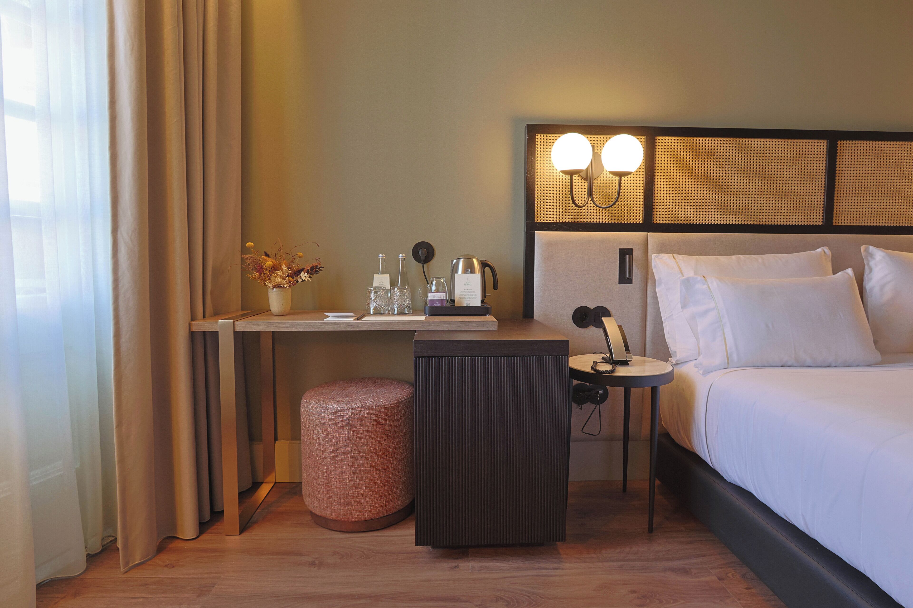 Een hotelkamer met een bed, nachtkastje, lamp en een klein krukje.