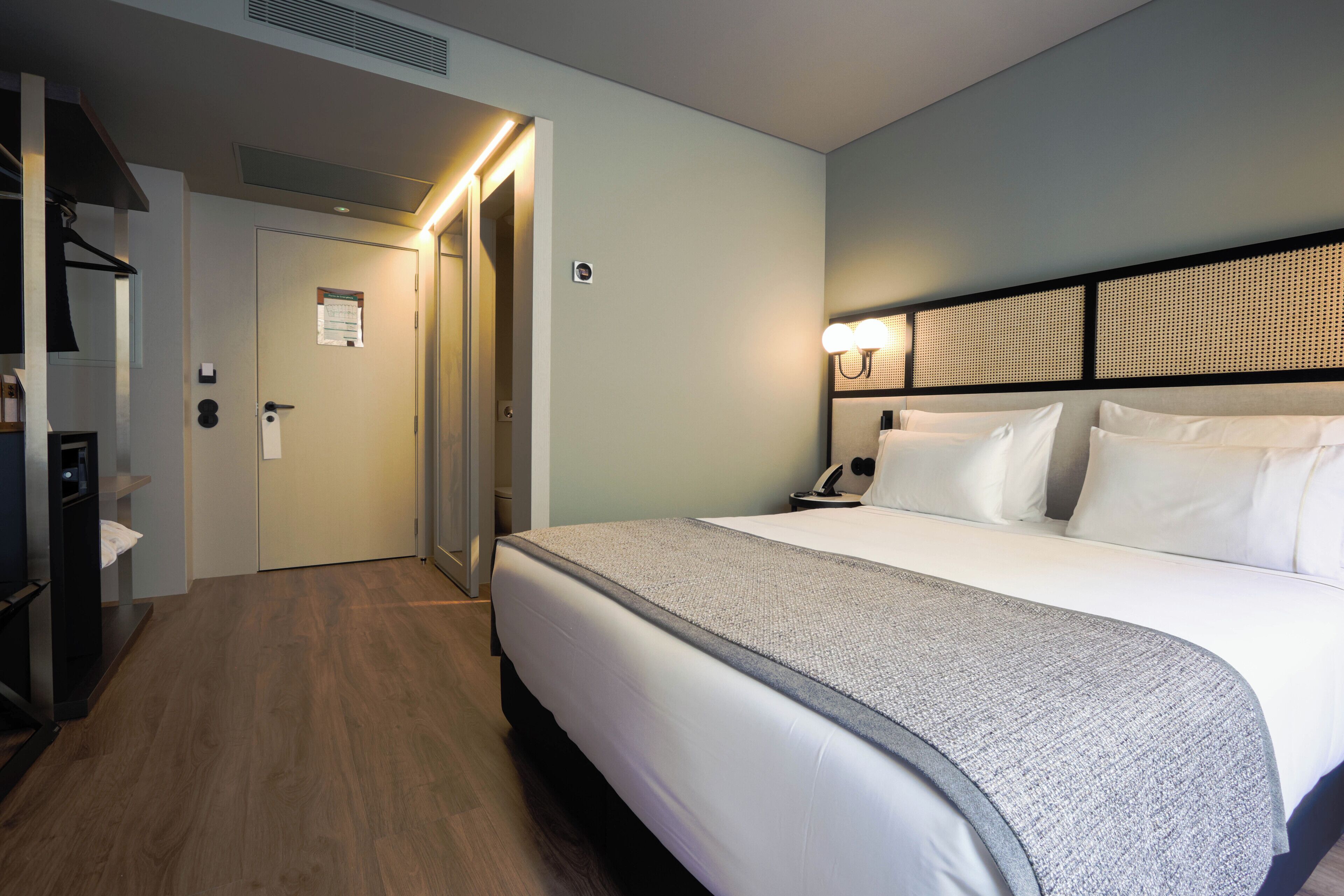A hotel room with a bed, bedside table, lamp, and a door leading to a hallway.
