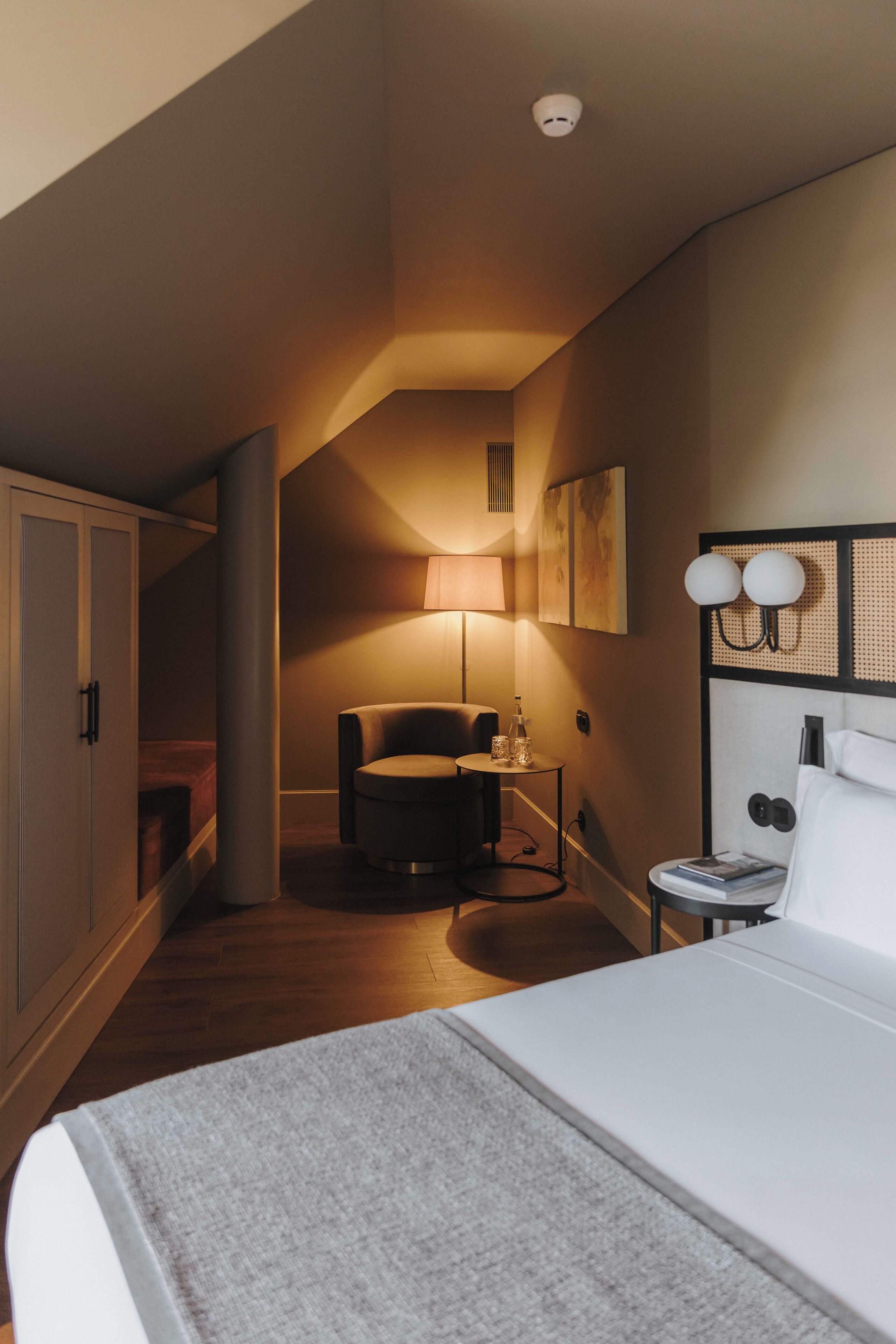A hotel room with a bed, a desk, a chair, a lamp, and a wall-mounted light fixture.
