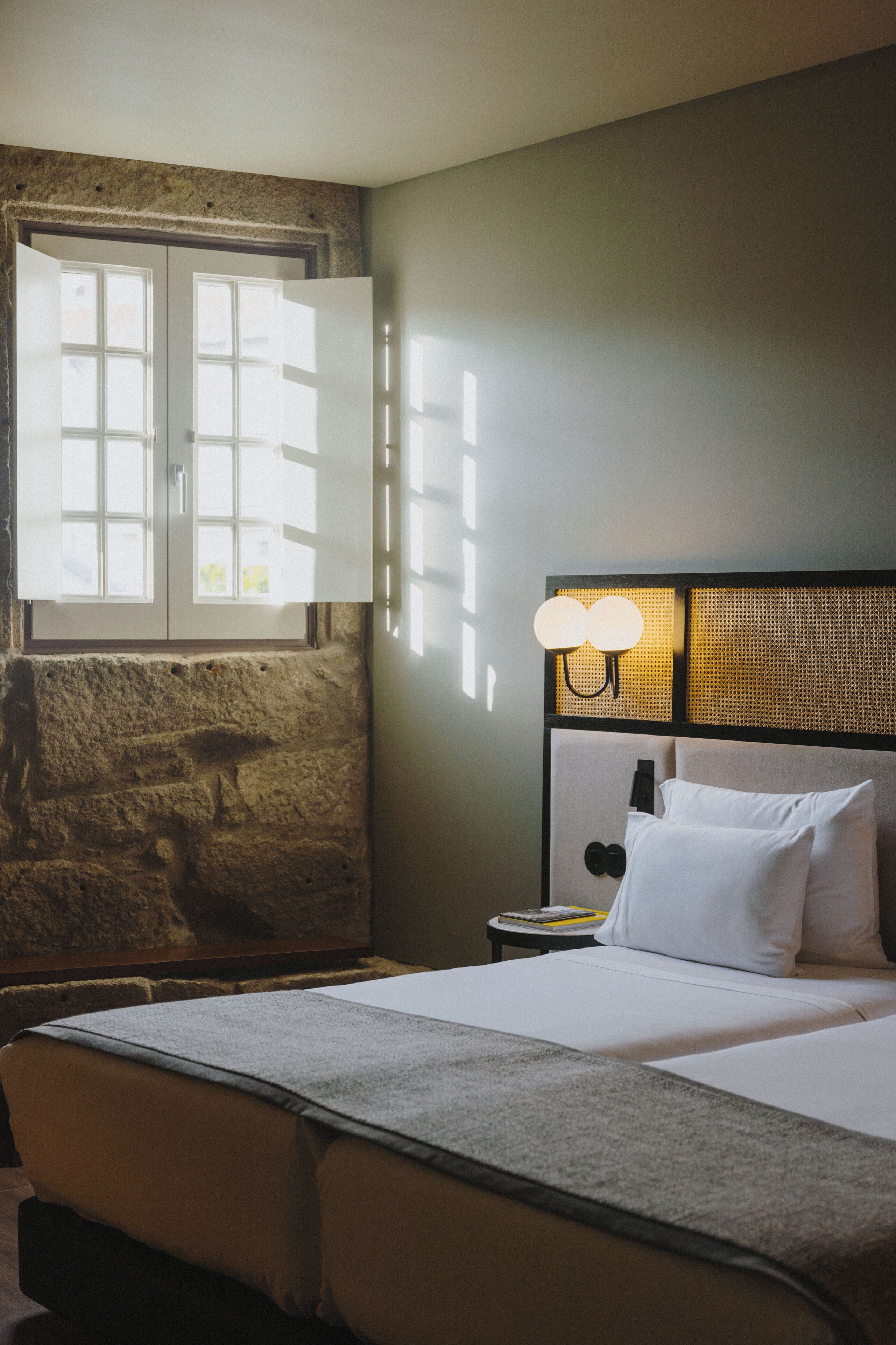 Una habitación de hotel con una cama, una mesita de noche, una lámpara y un ventanal con cortinas.