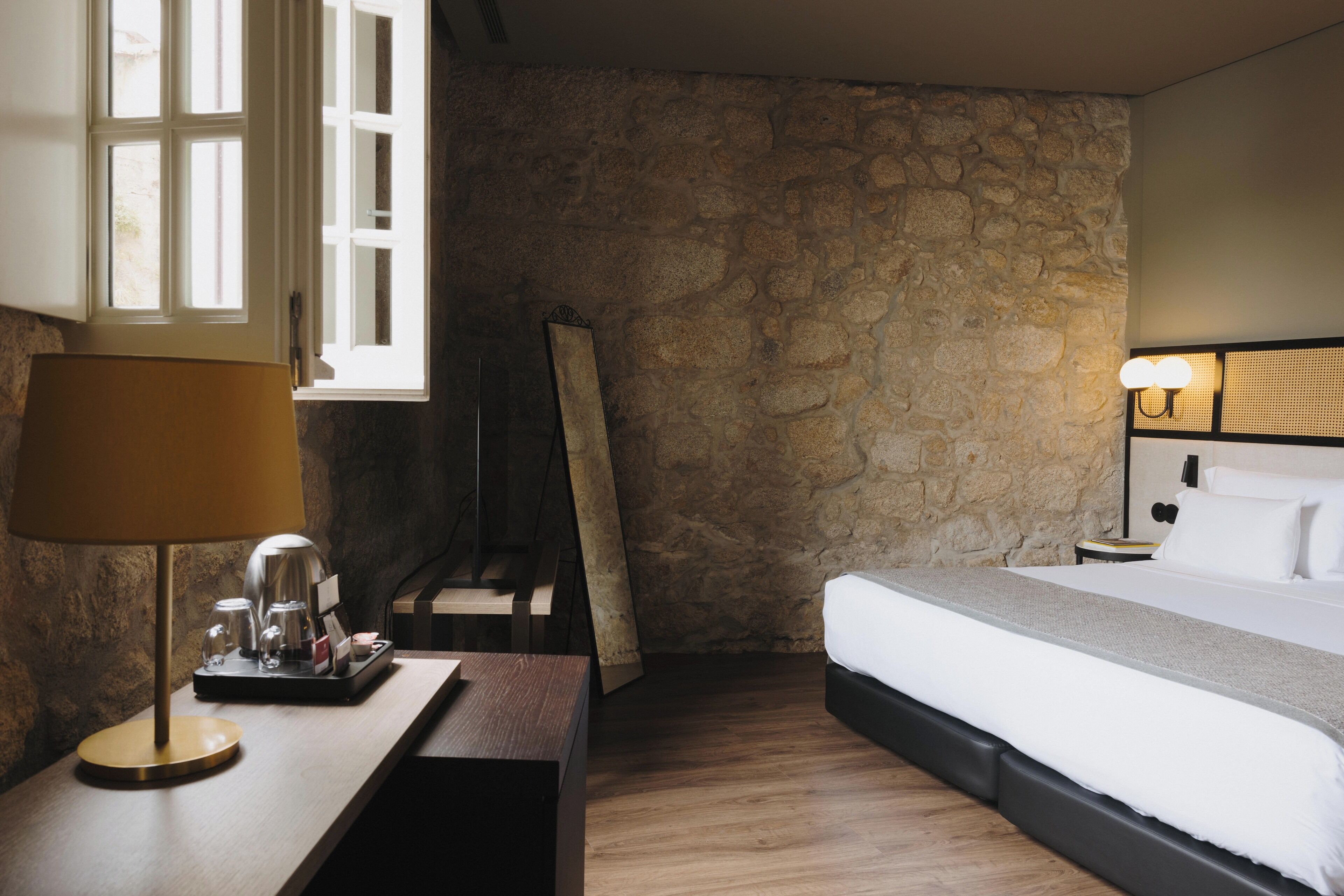 Quarto de hotel com parede de pedra, uma cama, mesa de cabeceira com abajur e uma janela.