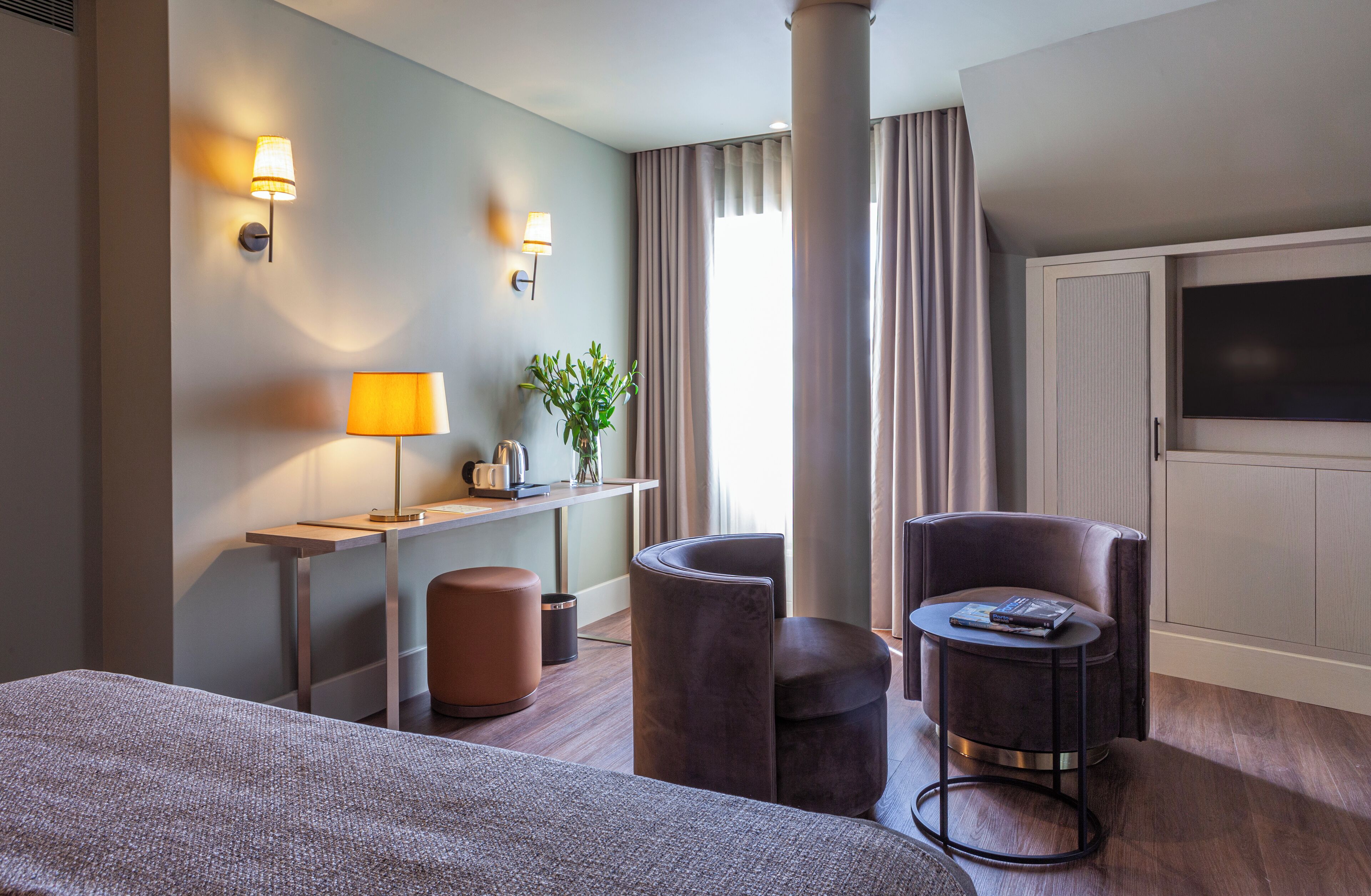 A hotel room with a bed, a desk with a lamp, a chair, a small table, and a TV.