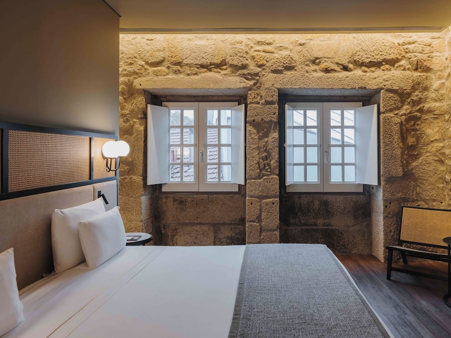Une chambre avec un mur en pierre, un lit avec une literie blanche, deux fenêtres, une table de chevet avec une lampe et une petite table ronde avec une plante.