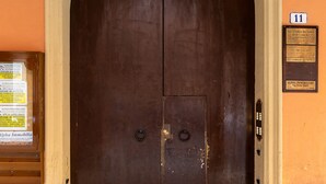 Uma porta de madeira escura e grande com um arco bege, uma maçaneta de metal e uma pequena placa na parede à direita.