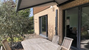 A wooden deck with a table and chairs, a brick building with a sliding glass door, and a tree in the background. - Modern country house with spacious rooms and swimming pool (Doingt)