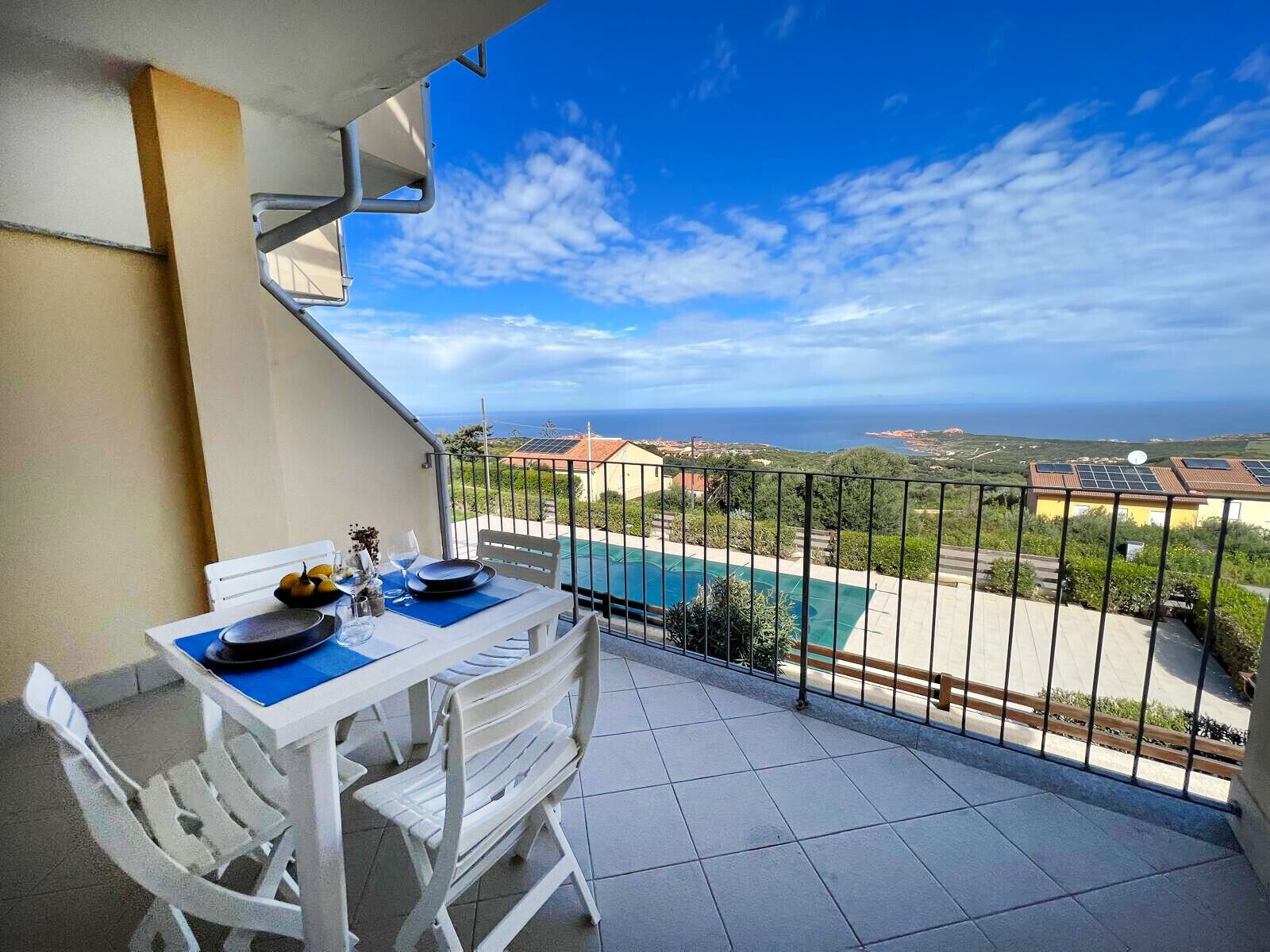 Un balcon avec une table dressée pour deux personnes, offrant une vue sur une piscine et un panorama pittoresque de la mer et des bâtiments éloignés.