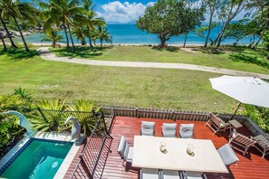 Una piscina con cubierta de madera, mobiliario exterior y vista a la playa.