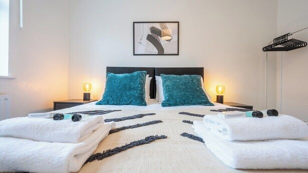 Ein ordentlich bezogenes Bett mit zwei Kissen, einer Nachttischlampe und einem gerahmten Kunstwerk an der Wand.