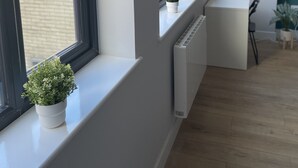 Ein modernes Zimmer mit großen Fenstern, einem Heizkörper, einem Schreibtisch und einer Wanduhr.
