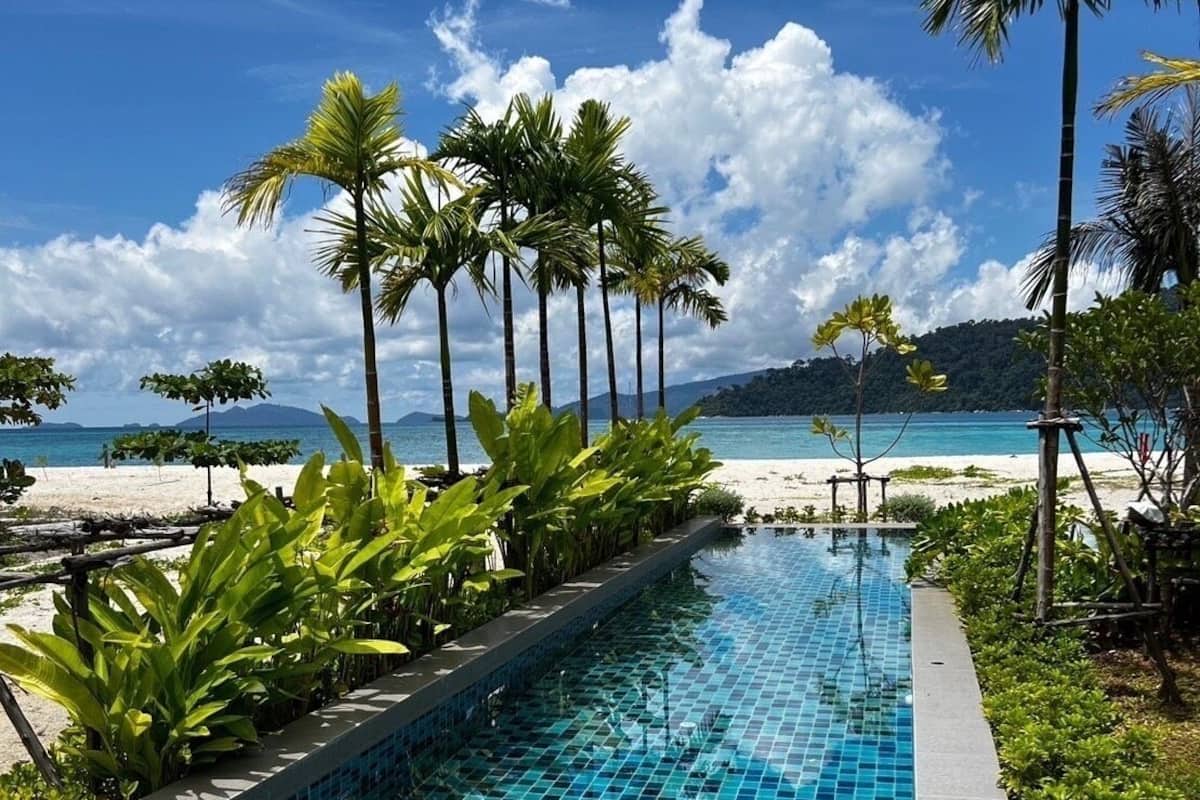 A swimming pool with blue tiles, surrounded by palm trees and a beach in the background.