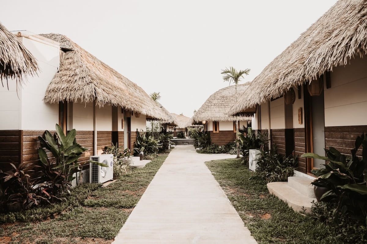 A pathway leading to thatched-roof cottages with a minimalist design, surrounded by tropical plants.