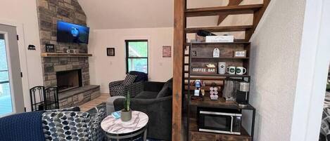 A living room with a stone fireplace, a television, a coffee bar, and a staircase.