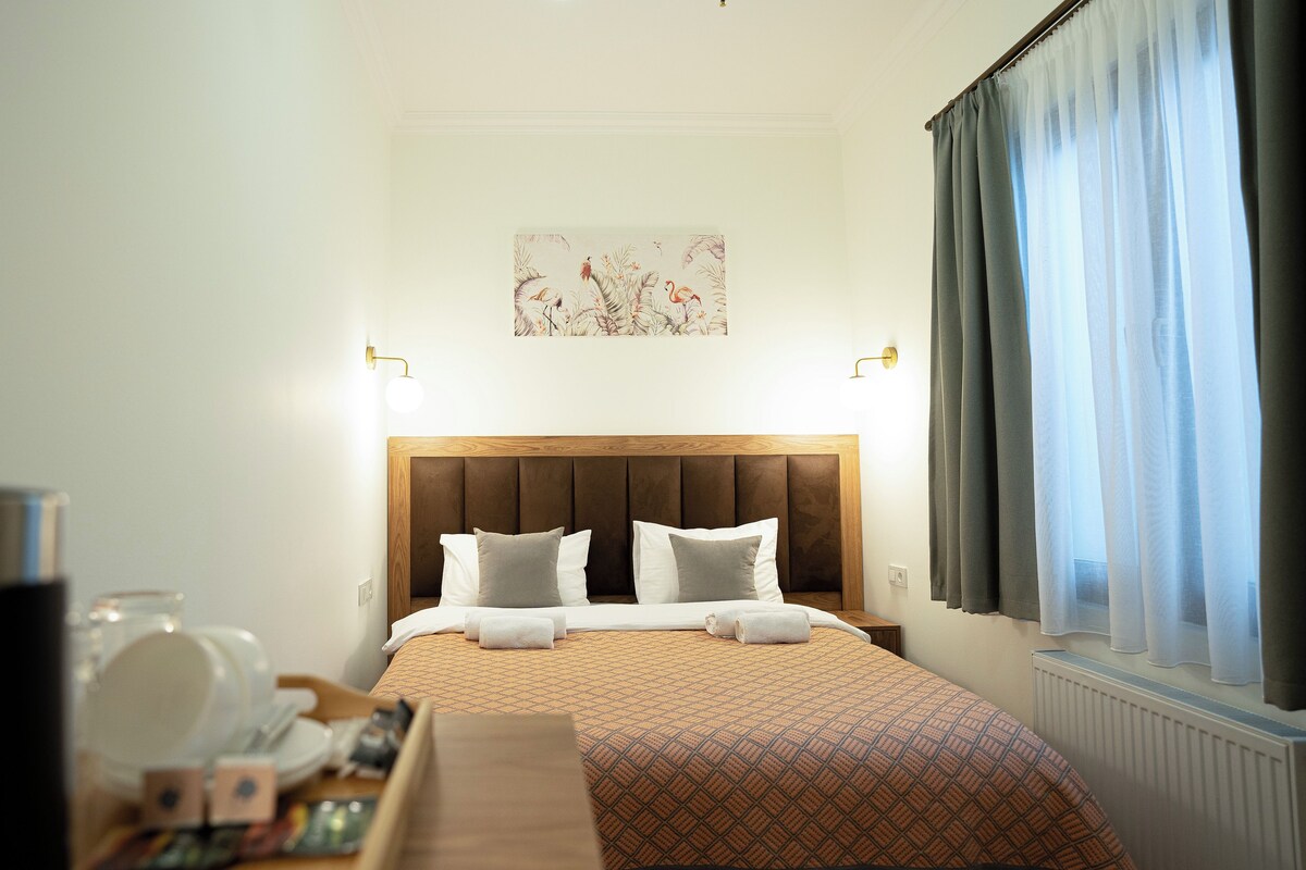 A hotel room with a bed, a headboard, a painting, a window with curtains, and a table with items.