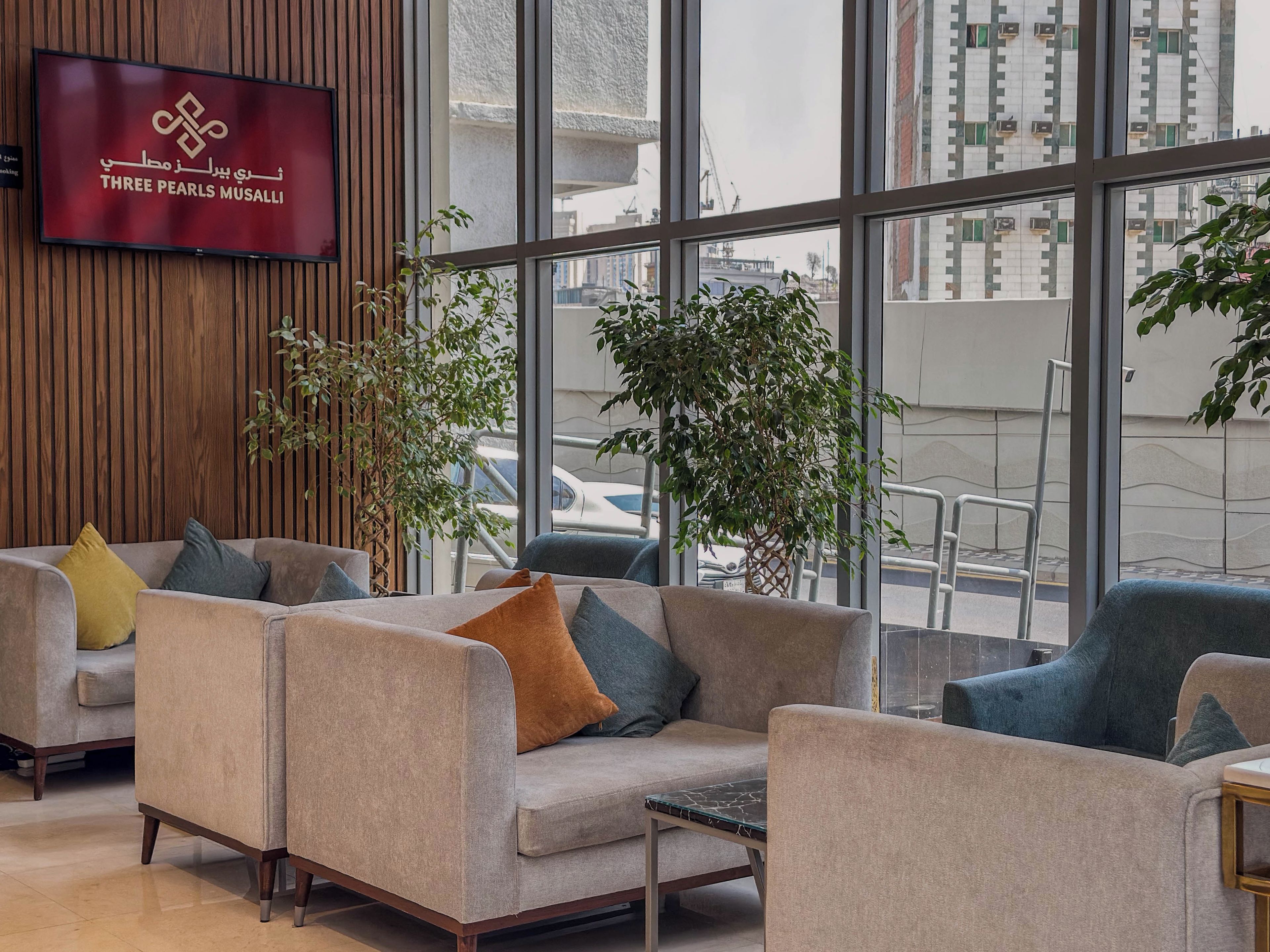 A hotel lobby with a large TV screen displaying the hotel name, multiple seating areas with cushions, and large windows offering a view of the cityscape.