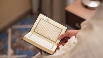 Een persoon houdt een boek met Arabische tekst vast, vermoedelijk een religieuze tekst, in een kamer met een kruis aan de muur en een klein tafeltje met een kom.