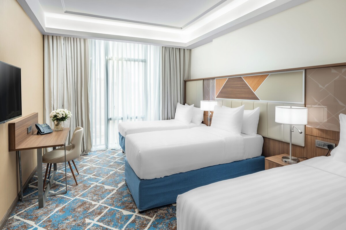 Une chambre d’hôtel avec deux lits, un bureau avec une chaise, une télévision et une grande fenêtre avec des rideaux.