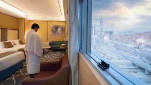 Quarto de hotel com uma grande janela que oferece vista para a cidade, uma cama, um sofá e uma pequena mesa com um vaso.