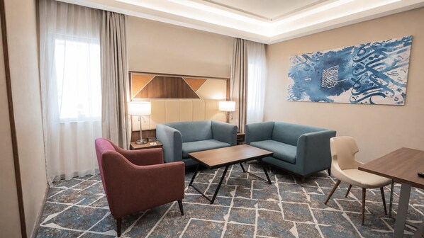 Ein Hotelzimmer mit einer Sofagarnitur, einem kleinen Tisch, einem Sessel, einer Lampe und einer Wand mit einem blauen abstrakten Gemälde.