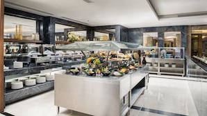 Uma área de buffet com diversos alimentos, incluindo frutas, legumes e pratos prontos, dispostos em um balcão de mármore com vitrines de vidro.
