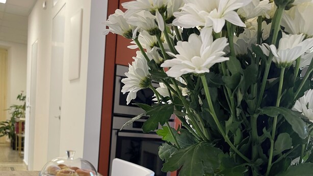 Een eettafel gedekt met witte borden, bestek en een glas water, met een vaas witte bloemen en een potje jam erop.