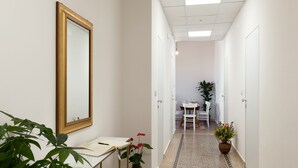 Een hal met een witte muur, een spiegel met gouden lijst, een wit bureau met een notitieboek, een potplant en een zithoek met een tafel en stoelen.
