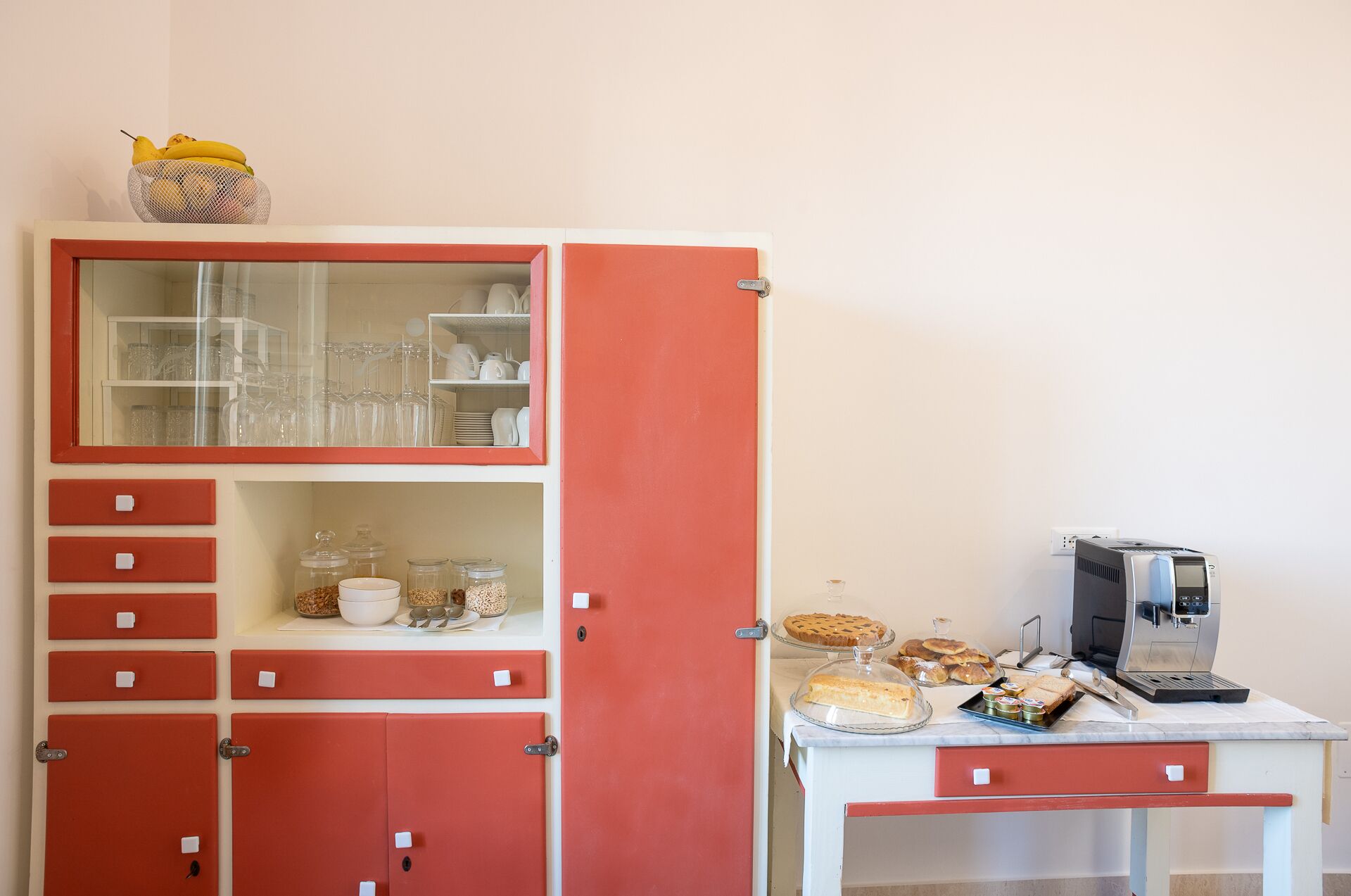 Une petite cuisine équipée d’une machine à café, d’une petite table avec des aliments et d’un panier de fruits posé sur le comptoir.