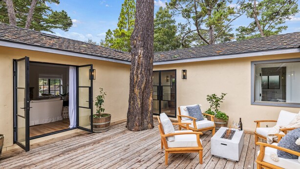 Una terraza de madera con mobiliario exterior, un árbol grande y un edificio con tejado de tejas.