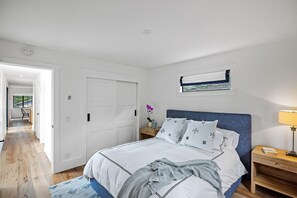 Ein Schlafzimmer mit Bett, Nachttisch, Fenster mit Jalousien und einer Tür zu einem anderen Raum.