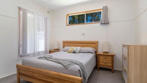 Ein Schlafzimmer mit einem hölzernen Bett, einem Nachttisch mit Lampe, einer Kommode und einem Fenster mit Blick auf Bäume.