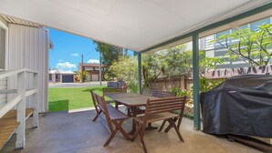 Eine überdachte Terrasse mit Tisch und Stühlen, einem Grill und Blick auf eine Wohngegend mit Haus und Bäumen.
