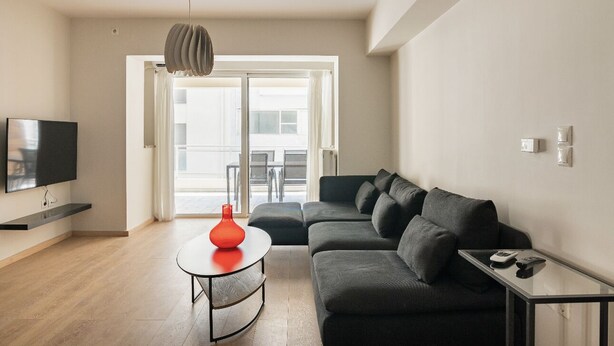 Un salon moderne avec un canapé gris, une table basse ronde surmontée d’un vase rouge, un téléviseur à écran plat et un luminaire suspendu.