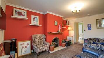 Un salon aux murs rouges, avec une cheminée, un fauteuil à motifs, un canapé en cuir et des œuvres d’art encadrées accrochées aux murs.