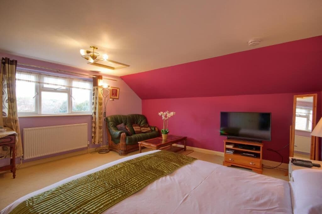 Un dormitorio con una cama grande, un sillón verde, una televisión y un ventanal con cortinas.