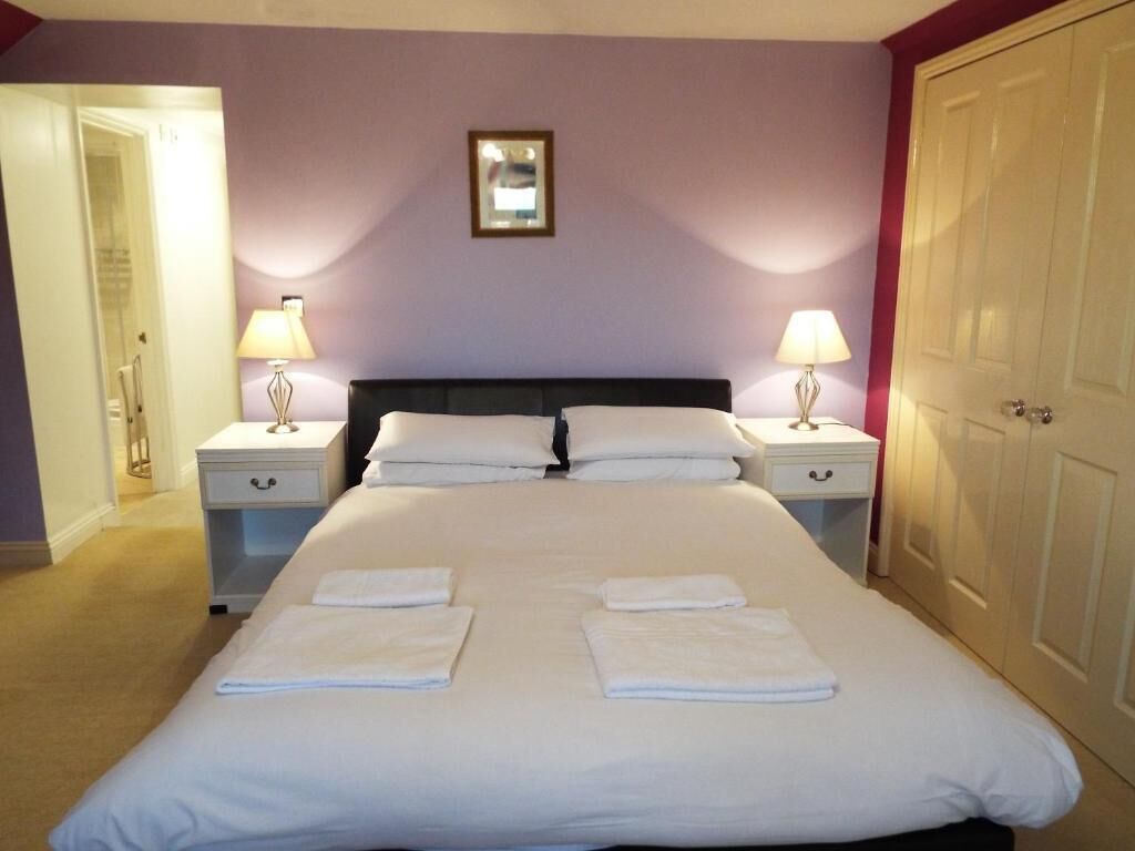 A neatly made bed with white linens and towels, two bedside tables with lamps, a framed picture on the wall, and a door to the right.