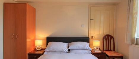 Una habitación de hotel con una cama, dos mesitas de noche, un armario de madera, una puerta y un ventanal con cortinas.