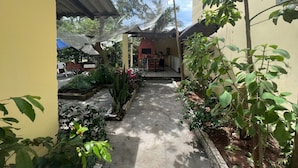 Un patio con zona de estar cubierta, rodeado de plantas y un pequeño edificio.