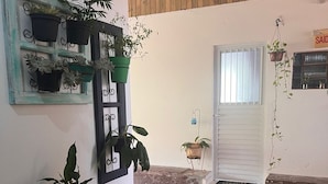 Una habitación con piso de baldosas, techo de madera, puerta blanca y una ventana con barrotes. Hay plantas en macetas sobre una repisa y un taburete amarillo.