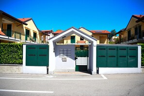 A gated entrance to a residential area with a white building featuring a triangular roof and green metal gates.
