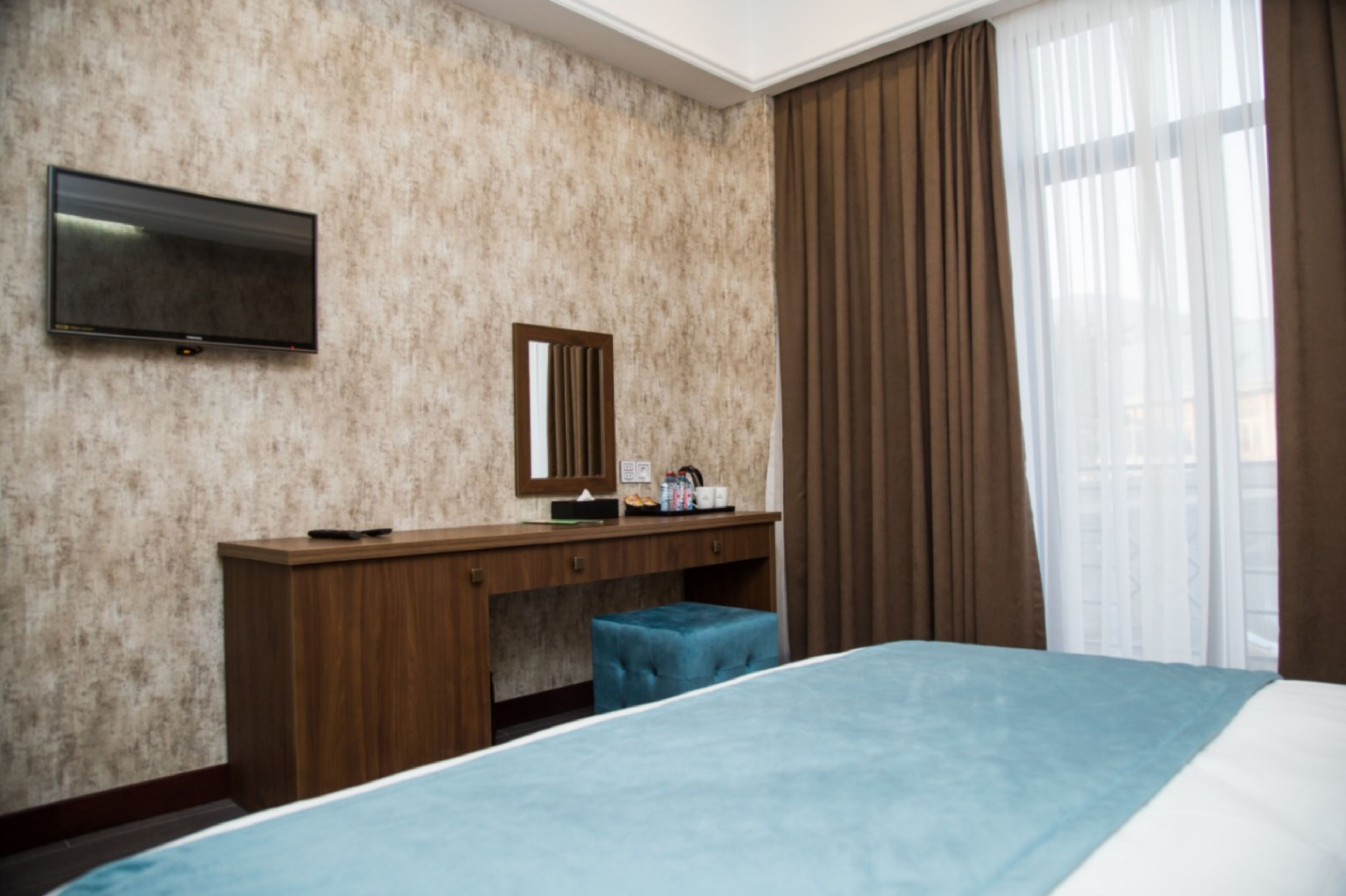 Una habitación de hotel con una cama, un televisor montado en la pared, un escritorio de madera con un espejo y una ventana con cortinas.