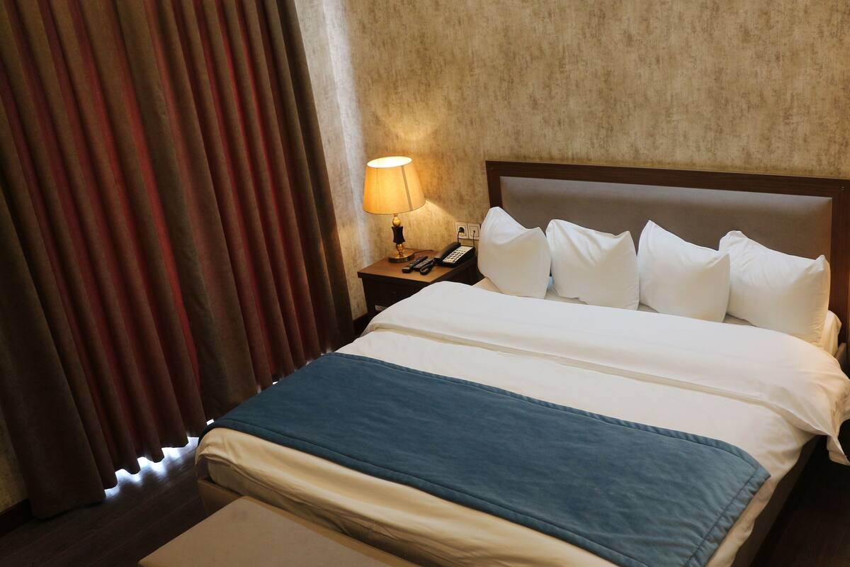 A hotel room with a large bed, two bedside lamps, a nightstand with a phone and remote controls, and a window with curtains.