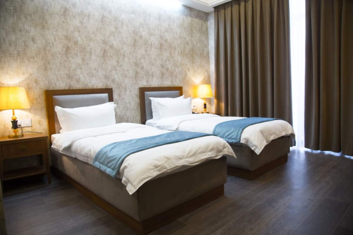 A hotel room with two beds, a wooden headboard, a nightstand with a lamp, and a large window with curtains.