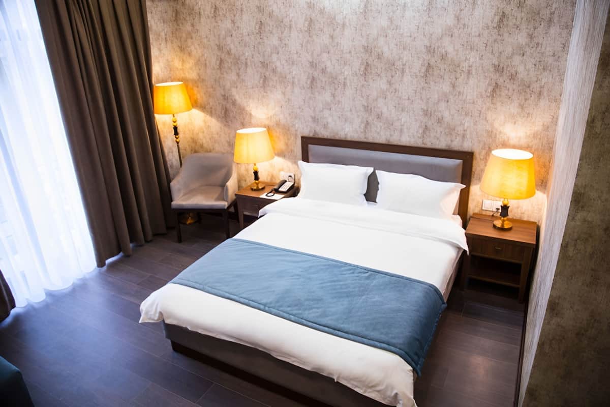 A hotel room with a large bed, two bedside tables with lamps, a chair, and a view of the outdoors through curtains.