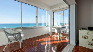 Amplia habitación con grandes ventanales con vistas al mar, un comedor con sillas blancas y un armario blanco con detalles decorativos.
