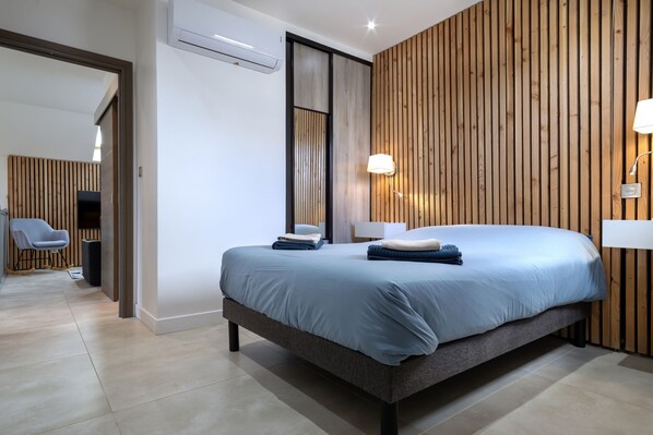 A modern hotel room with a large bed, wooden panel wall, and a seating area with a chair and table.